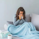 Donna raffreddata che si soffia il naso sul divano con coperta e bevanda calda