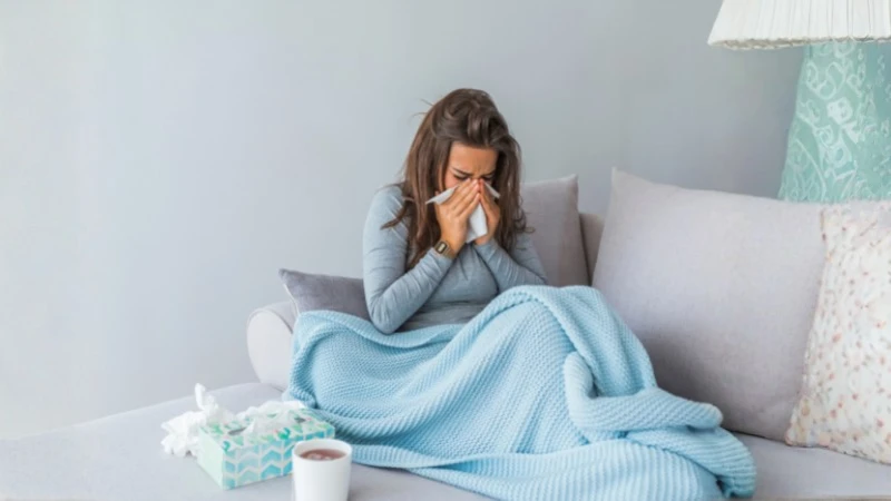 Donna raffreddata che si soffia il naso sul divano con coperta e bevanda calda